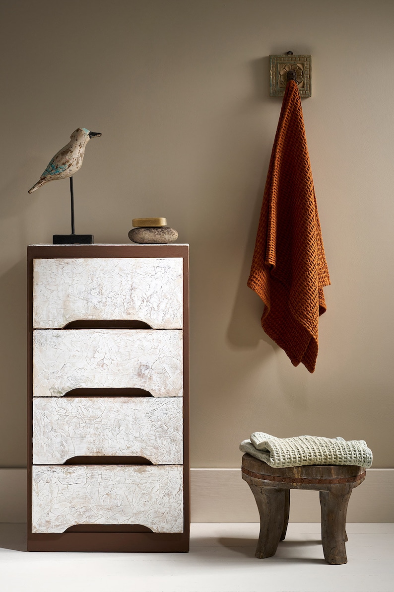 May include: A white dresser with four drawers and brown trim sits on a white floor. A small wooden stool with a woven cushion sits in front of the dresser. A brown towel hangs on a hook on the wall. A small wooden bird figurine sits on top of the dresser.