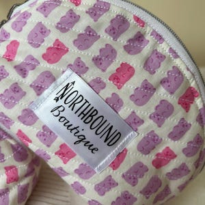 May include: A white quilted makeup bag with a purple and pink gummy bear print. The bag has a zipper closure and a white label that says "Northbound Boutique".
