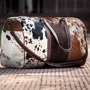 May include: A brown, white, and black cowhide duffel bag with a brown leather strap. The bag has a zipper closure and a top handle.