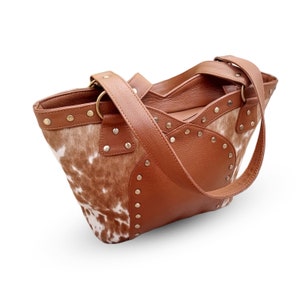 May include: A brown leather tote bag with a cowhide pattern and silver studs. The bag has two straps and a top zipper closure.