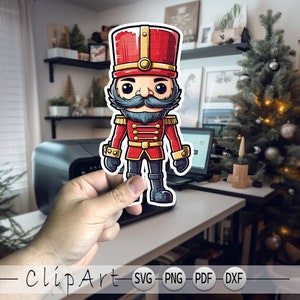 Może przedstawiać: Kreskówkowa ilustracja czerwonego i złotego żołnierzyka z czarnym zarostem. Żołnierzyk stoi z rękami opuszczonymi wzdłuż ciała i jest ubrany w czerwony mundur z złotymi ozdobami. Żołnierzyk trzyma w prawej ręce plastikową broń.