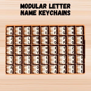 May include: A wooden box filled with white letter tiles, numbers, and symbols. The tiles are arranged in rows and columns, and each tile has a black letter or number printed on it. The box is labeled "Modular Letter Name Keychains".