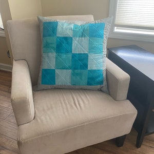 May include: A light gray armchair with a blue and gray patterned pillow. The pillow has a checkered design with a light blue background and darker blue squares.