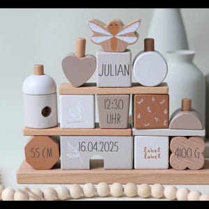 Könnte beinhalten: Ein Holz-Stapelspielzeug mit verschiedenen Formen und Blöcken, darunter ein Herz, ein Schmetterling, ein Kreis und ein Bienenstock. Die Blöcke sind mit Text verziert, darunter "Julian", "12:30 Uhr", "16.04.2025", "55 cm" und "4100 g".