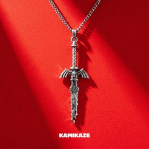 May include: A silver-toned metal sword pendant hanging from a matching chain. The sword features a detailed hilt with wing-like crossguards and a blade with an intricate, textured pattern. The word KAMIKAZE is printed in white at the bottom.