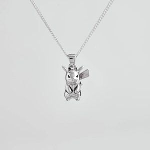 May include: A silver-toned metal pendant shaped like a stylised creature with pointed ears and a lightning bolt-shaped tail, suspended from a delicate silver-toned chain. The pendant features a smooth, reflective surface with engraved facial details.