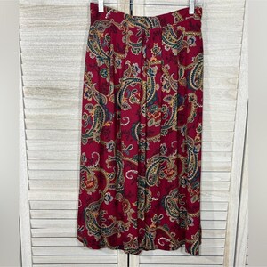 May include: A pair of wide-leg trousers featuring a deep red base color with a multi-colored paisley pattern in shades of blue, gold, and cream. The trousers have a high-waisted design with a structured waistband and a flowing, loose silhouette.