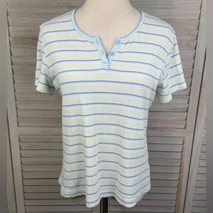 May include: A short-sleeved, striped shirt in light blue, yellow, and white, displayed on a mannequin. The shirt has a henley neckline with three light blue buttons. The fabric appears to be ribbed.