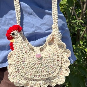 May include: Cream-colored crochet shoulder bag with a chicken design. The bag features a red comb and yellow accents, a pink button closure, and a scalloped edge. The strap is also crocheted.
