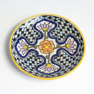 May include: A round ceramic plate with a yellow rim and a white background. The plate features a blue and black checkered pattern with a floral design in the center. The floral design includes four purple flowers with yellow centers and green stems, and a large orange flower with yellow petals in the center.