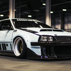 May include: A white custom-built car with black accents and a large rear wing. The car has a wide body kit and large wheels. The car is parked in a garage.