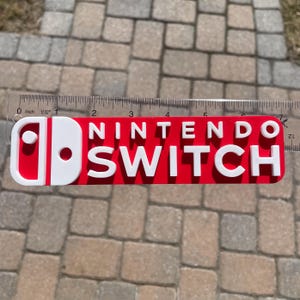 May include: A red and white Nintendo Switch logo, approximately 15 cm long, is displayed against a brick background. The logo features the Nintendo Switch branding in white, with the iconic controller design on the left.