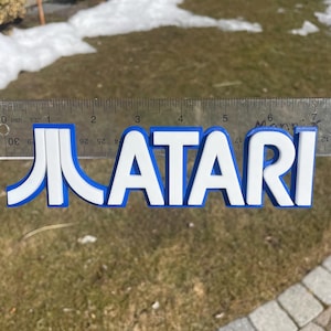 May include: A white 3D printed Atari logo with blue outlines. The logo is approximately 15 cm wide and is measured against a ruler.