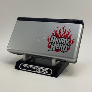 May include: A silver Nintendo DS handheld game console with a "Guitar Hero" sticker in red and black flames. The console is on a black stand with "Nintendo DS" printed in white.