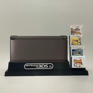 May include: A black Nintendo 3DS XL console stand with a white label that reads "NINTENDO 3DS XL". The stand has a slot for the console and a shelf for five game cartridges. The cartridges are for the games "Super Smash Bros. for Nintendo 3DS", "The Legend of Zelda: A Link Between Worlds", "Pokemon X", and "Mario Kart 7".