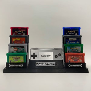 以下が含まれることがあります： ゲームボーイマイクロ用の黒いスタンドで、2列のゲームカートリッジが置かれています。 上段には、ポケモン ルビー、クラッシュ スーパーパック、ニックトゥーンズ ビデオコレクションが置かれています。 下段には、ポケモン エメラルド、ポケモン ファイアレッド、青いカートリッジに「PAC-MAN WORLD 2」と書いてあります。 スタンドには、「GAME BOY Micro」と書いてあります。