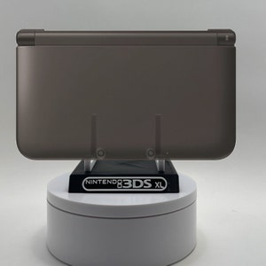 May include: A dark gray Nintendo 3DS XL handheld gaming console on a white display stand. The console is closed and the Nintendo 3DS XL logo is visible on the stand.