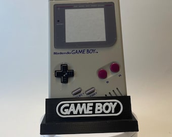 Puede incluir: Una consola de videojuegos portátil Nintendo Game Boy gris con una pantalla en blanco y negro, un pad direccional negro y dos botones morados. La Game Boy está sentada en un soporte negro con las palabras "GAME BOY" en letras blancas.
