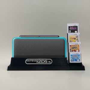 May include: A black stand with the text "NINTENDO 2DS XL" holds a teal and black Nintendo 2DS XL console. The stand also holds a stack of five Nintendo 3DS game cartridges, including "The Legend of Zelda: A Link Between Worlds", "The Legend of Zelda: Ocarina of Time 3D", "Pokemon Sun", "Pokemon Moon", and "Pokemon Alpha Sapphire".