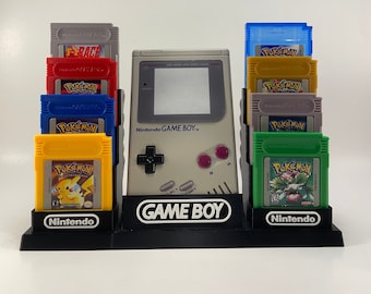 Puede incluir: Un soporte negro con una consola Game Boy Color gris y ocho cartuchos de juego. Los cartuchos son todos juegos de Pokemon, con títulos como "Pokemon Red" y "Pokemon Yellow".