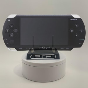 May include: A black Sony PlayStation Portable (PSP) handheld gaming console with a white trim and a black stand. The PSP is on a white circular base.