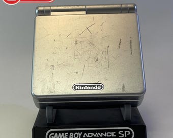 May include: A silver Game Boy Advance SP handheld gaming console with a black stand. The console has a scratched surface and the Nintendo logo is visible on the front.