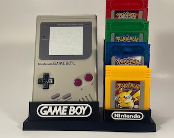 Puede incluir: Un soporte negro con una consola portátil Game Boy gris y cinco cartuchos de juego Pokemon de colores. El soporte tiene las palabras "GAME BOY" impresas en la parte delantera y "Nintendo" impresas en la parte inferior.