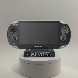 以下が含まれることがあります： 黒とグレーのPlayStation Vita携帯ゲーム機がスタンドに置かれています。コンソールには大きな画面、コントロールボタンがあり、上部左側に「SONY」の文字があります。スタンドには「PS VITA」の文字があります。