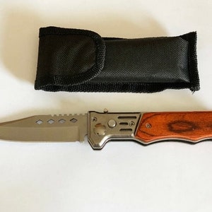 Könnte beinhalten: Ein silbernes Klappmesser mit einem Holzgriff und einer schwarzen Nylon-Scheide. Die Klinge hat eine gezahnte Kante und ein kleines Loch für eine Schnur.