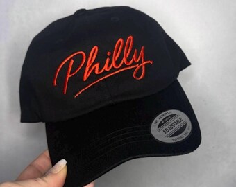 Puede incluir: Gorra de béisbol negra con la palabra "Philly" bordada en escritura naranja. La gorra tiene una visera curva y una correa ajustable. Una pegatina plateada con texto está en la visera.