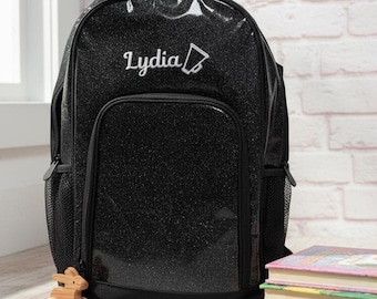 Puede incluir: Mochila negra con purpurina con el nombre "Lydia" bordado en blanco. La mochila tiene un bolsillo frontal, bolsillos laterales de malla y un asa superior. Se ve un pequeño juguete de madera y una pila de libros.