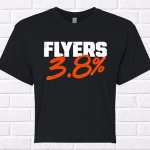 Puede incluir: Camiseta negra con la palabra "FLYERS" en letras blancas y el n&uacute;mero "3.8%" en naranja. La camiseta se muestra sobre un fondo de pared de ladrillo blanco.