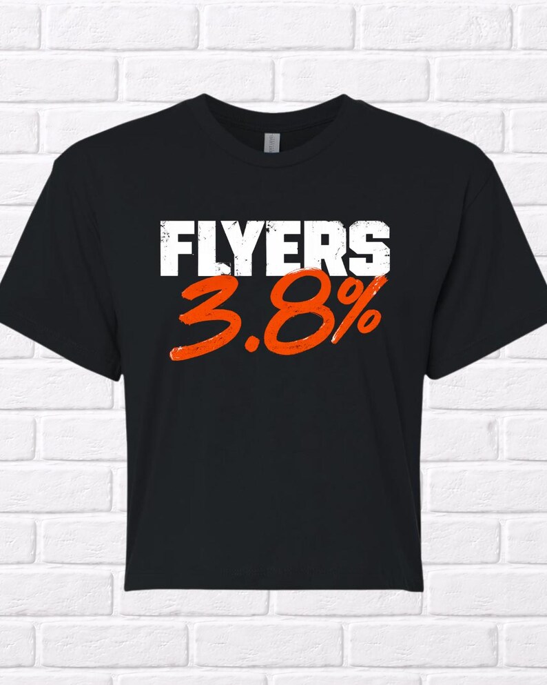 Puede incluir: Camiseta negra con la palabra "FLYERS" en letras blancas y el n&uacute;mero "3.8%" en naranja. La camiseta se muestra sobre un fondo de pared de ladrillo blanco.