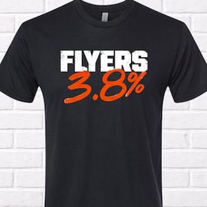 Puede incluir: Camiseta negra con la palabra "FLYERS" en blanco sobre los n&uacute;meros naranjas "3.8%". La camiseta tiene cuello redondo y mangas cortas. El fondo es una pared de ladrillos blancos.