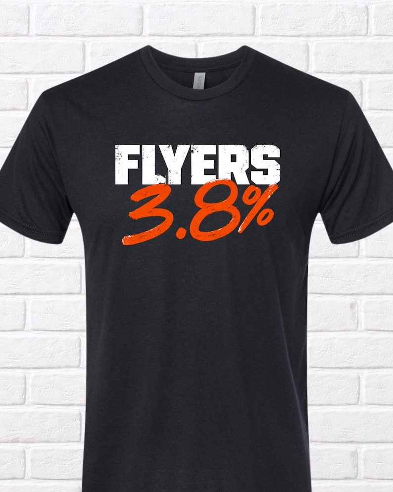 Puede incluir: Camiseta negra con la palabra "FLYERS" en blanco sobre los n&uacute;meros naranjas "3.8%". La camiseta tiene cuello redondo y mangas cortas. El fondo es una pared de ladrillos blancos.