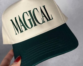 Puede incluir: Gorra de béisbol con corona color crema y visera verde oscuro. La palabra "MAGICAL" está bordada en verde oscuro en la parte delantera de la gorra. La gorra está siendo sostenida por una persona.