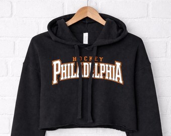 Puede incluir: Sudadera corta negra con la palabra "PHILADELPHIA" en letras blancas con contorno naranja, y la palabra "HOCKEY" encima en letras naranjas. La sudadera está colgada en una percha de madera contra una pared de ladrillos blancos.