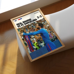 May include: Framed poster featuring a soccer match between Real Madrid and FC Barcelona, with a score of 2-3. The image shows a player holding up a jersey. The poster has a wooden frame and is displayed on a wooden surface.