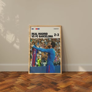 May include: A framed poster featuring a soccer player holding up a jersey. The poster has the text "REAL MADRID VS FC BARCELONA 2-3" with the date "17th May 2017" at the top. The poster is in a wooden frame.