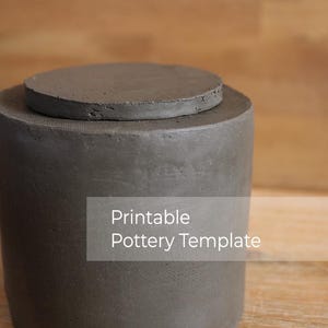 May include: A grey concrete pot with a circular lid, featuring the text "Printable Pottery Template". The pot has a textured finish and sits on a light brown wooden surface. The design is simple and modern.