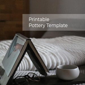 Könnte beinhalten: Ein offenes Buch, das auf einem dunkelgrauen Tablett steht, mit einer Brille und einer kleinen, hellgrauen Keramikschale. Der Text "Printable Pottery Template" wird auf dem Bild angezeigt. Die Szene spielt sich auf einem Bett mit einer weißen Decke ab.