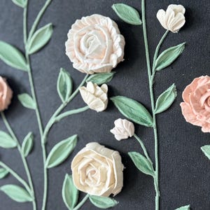 May include: A close-up of a floral design on a dark background. The design features raised, textured roses and buds in shades of cream and peach, with green leaves and stems. The artwork has a three-dimensional effect.