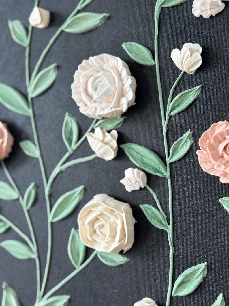 May include: A close-up of a floral design on a dark background. The design features raised, textured roses and buds in shades of cream and peach, with green leaves and stems. The artwork has a three-dimensional effect.