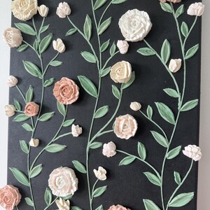 May include: A floral canvas artwork with textured roses in cream and peach hues, set against a black backdrop. The green leaves and stems add contrast. The raised design gives a three-dimensional effect, creating a tactile and visually appealing piece.