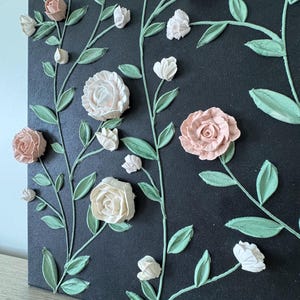 May include: A canvas artwork with textured floral designs. It features light pink, white, and peach-coloured roses and buds, with green leaves and stems against a dark background. The dimensional flowers create a textured, artistic effect.