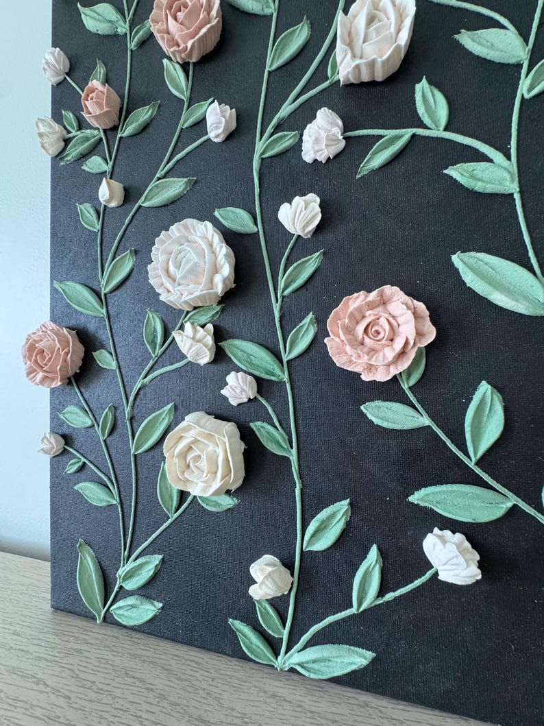 May include: A canvas artwork with textured floral designs. It features light pink, white, and peach-coloured roses and buds, with green leaves and stems against a dark background. The dimensional flowers create a textured, artistic effect.