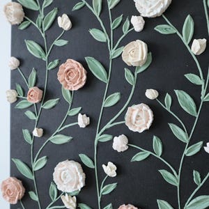 May include: A black canvas with a floral design. The artwork features raised, textured flowers in shades of white and pink, with green stems and leaves. The flowers are arranged in a repeating pattern.