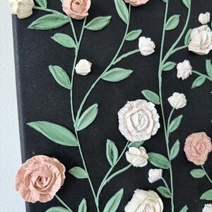 May include: A canvas artwork with raised floral details. The flowers are in shades of pink and white, with green leaves and stems against a black backdrop. The artwork has a textured appearance, with the flowers appearing to be 3D.
