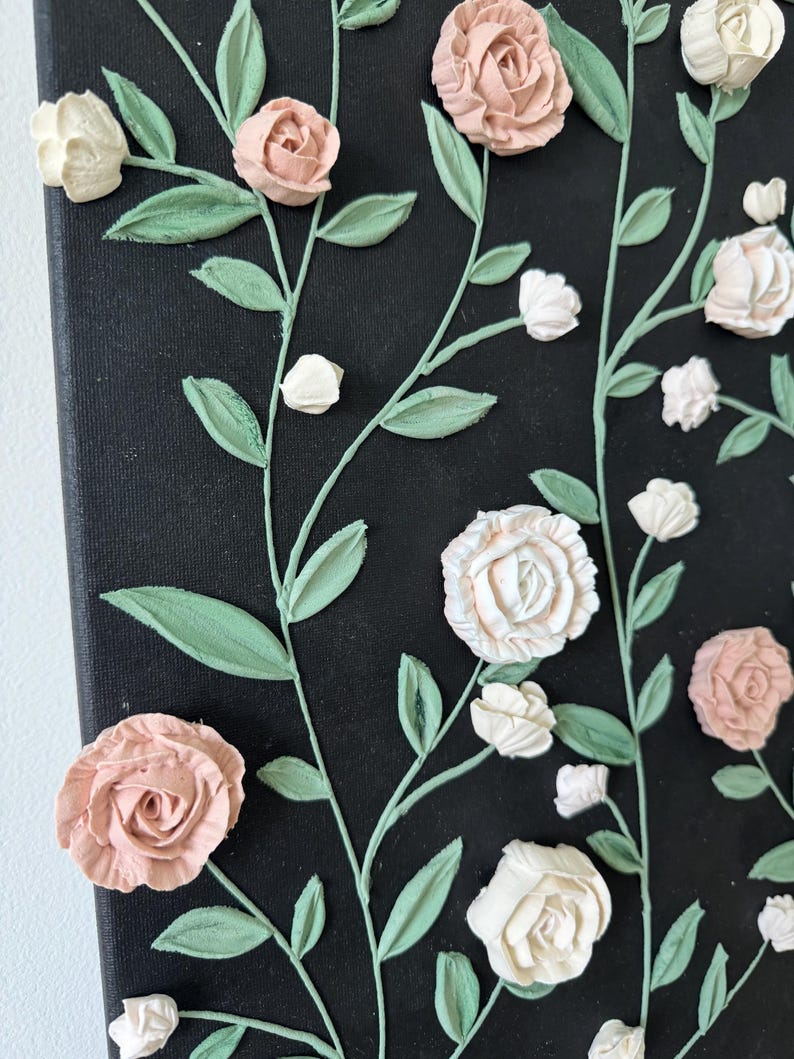 May include: A canvas artwork with raised floral details. The flowers are in shades of pink and white, with green leaves and stems against a black backdrop. The artwork has a textured appearance, with the flowers appearing to be 3D.