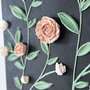 May include: Close-up of a canvas artwork with raised floral elements. The piece features peach roses and off-white buds, with green leaves and stems against a dark grey background. The textured design creates a three-dimensional effect.